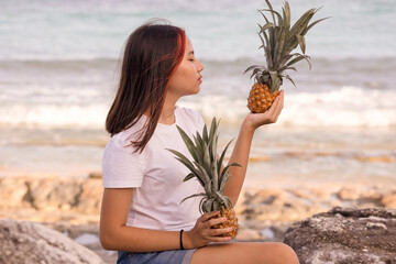 Teenager girl sitting on the stone, holding two whole pineapples. Profile of a face looking at pineapple. Organic fruit.
