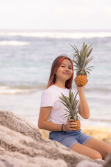 Happy teenager girl sitting on the stone, holding two whole pineapples. Smiling face. Organic fruit concept. Beach, Bali