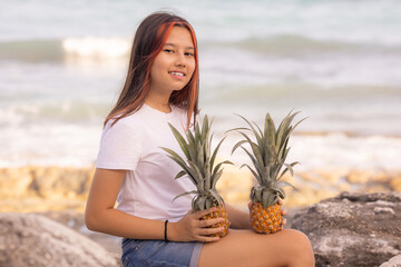Attractive teenager girl sitting on the stone, holding two whole pineapples. Smiling face. Organic fruit concept. Bali.
