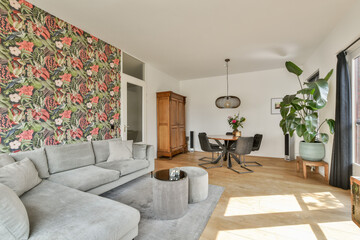 A stylish living room featuring elegant floral wallpaper, a cozy gray sofa, a wooden cabinet, and bright natural light. The design blends comfort with modern aesthetics.