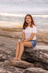 Charming teenager girl sitting on the stone on the beach. Summer vacation in Asia. Enjoy traveling to Bali island.