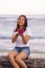 Teenager girl sitting on the stone, holding half a dragon fruit in front of her. Smiling face. Tropical fruit concept.