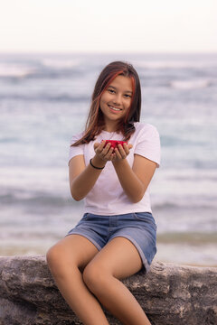 Teenager girl sitting on the stone, holding half a dragon fruit in front of her. Smiling face. Tropical fruit concept.