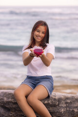 Girl sitting on the stone, holding half a dragon fruit in front of her. Selected focus on red dragon fruit. Blurred face