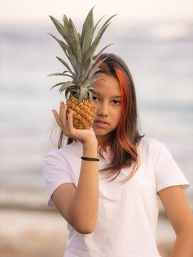 Teenager girl holding pineapple in front of her face. One eye closed by pineapple. Tropical fruit concept. Lifestyle.
