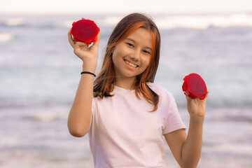 Portrait of charming Asian teenager girl holding red dragon fruit halves. Outdoor photoshoot with dragon fruits. Bali.