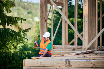 Carpenter constructing two-story wooden frame house near the forest. Bearded man wearing glasses hammering nails into structure. Concept of modern ecological construction.