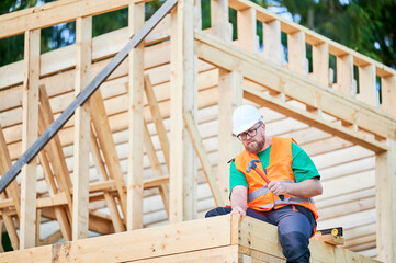 Fototapeta premium Carpenter constructing two-story wooden frame house near the forest. Bearded man wearing glasses hammering nails into structure. Concept of modern ecological construction.