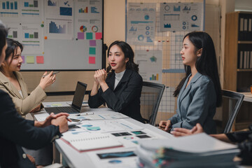 Asian businesswomen discussing and brainstorming, using laptop and financial documents, analyzing sales charts, working together on project strategy in boardroom at night