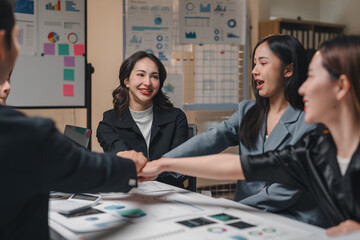Group of happy and joyful asian businesspeople joining hands together, celebrating their success in the office after a productive meeting analyzing financial charts and graphs