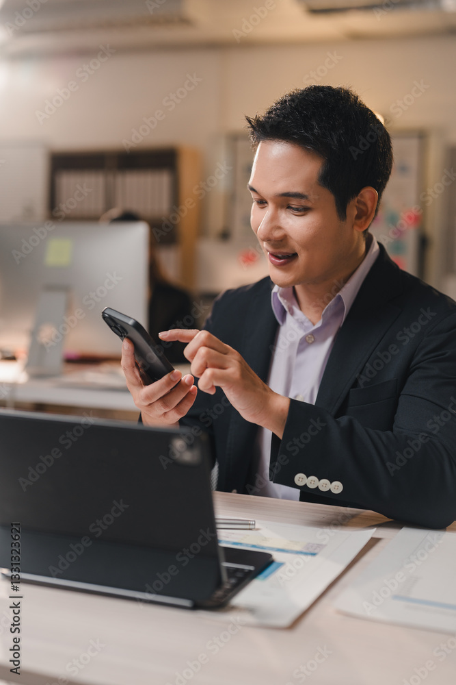Poster young, happy manager sitting at a desk in a modern office workspace, using a mobile phone and smilin