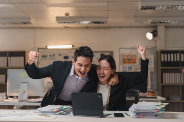Two overjoyed business people are celebrating success while working on a laptop in the office, raising their arms in excitement and laughter
