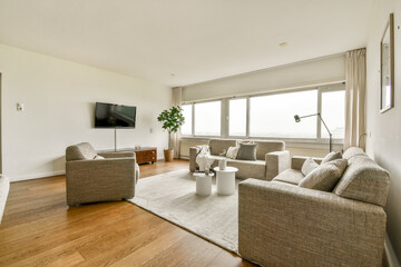 A spacious modern living room featuring light-colored walls, comfortable seating, a TV unit, and large windows that allow natural light to fill the space.