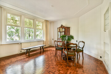 A spacious dining room featuring wooden flooring, large windows, vintage furniture, and a lush plant, creating a bright and inviting atmosphere.