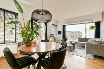 A contemporary living area featuring a round wooden table, stylish chairs, and large windows providing ample natural light and greenery.