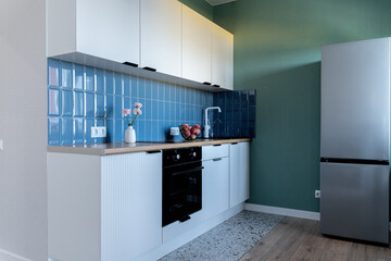 design of modern small kitchen. built-in kitchen with white cabinets, blue tile backsplash, wooden countertop. white kitchen unit. steel refrigerator, oven, flowers on vase, white faucet