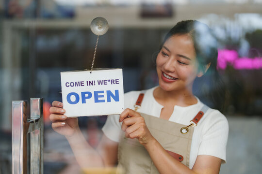 Young asian female shopkeeper smiling while hanging a come in! we're open sign on a glass door, showing her happiness for the reopening of the business after lockdown