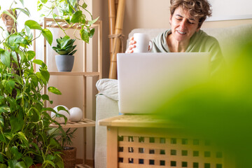Smiling mature freelancer working on a laptop while sipping coffee on a sofa in a cozy home office,...