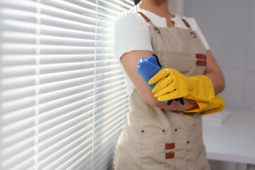 Professional cleaning lady wearing yellow gloves and holding a blue duster is standing next to a window with blinds, ready to start cleaning