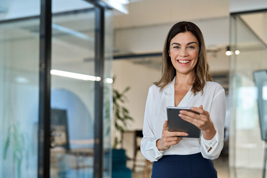 Smiling mature business woman executive, happy middle aged businesswoman entrepreneur, 45 years old company hr holding digital tablet looking at camera standing in office at work. Portrait.