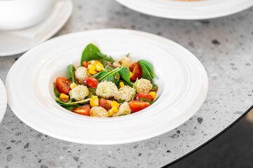 A fresh salad featuring spinach, cherry tomatoes, sweet corn, and sesame-coated balls, served in a clean, white dish on a textured concrete tabletop