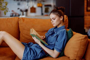 Young woman in denim dress reading book while sitting on the couch in cozy living room