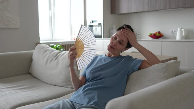 Unwell exhausted mature woman sitting on home couch waving paper fan to cool down. Overheated female suffers from stuffiness, feeling hot during heatwave in apartment without air conditioning. 