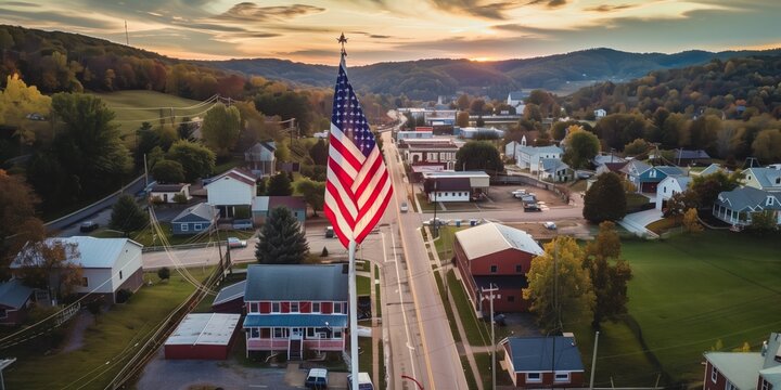Aerial view of a small American town with the United States flag in the foreground, surrounded by autumn foliage, rolling hills, and a serene sunset in the background