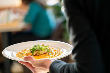 A close-up of a person holding a plate of gourmet food, featuring a salmon fillet on a bed of vibrant sauce, garnished with fresh herbs, in a lively dining atmosphere.