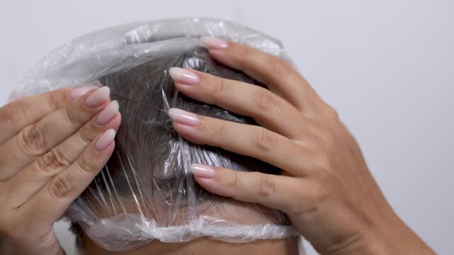 A young woman puts on a shower cap in a close-up shot to protect her hair from getting wet. Her hands carefully adjust the cap focusing on preparation for showering and hair care on a light background