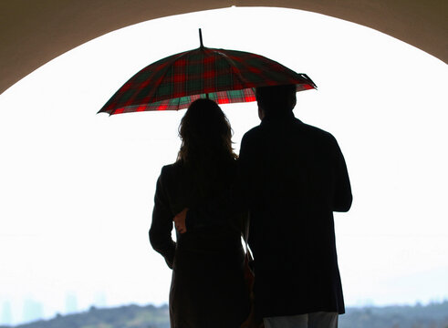 Hombre y mujer juntos bajo un arco de arquitectura con un mismo paraguas de color rojo y verde mirando el horizonte en un d&iacute;a de nubes, lluvia y tormenta en Madrid