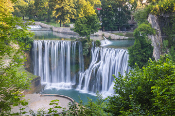 The Pliva Waterfall in Jajce town, Bosnia and Herzegovina, Europe. Majestic waterfall cascades down rocks into a pool, framed by lush green trees, creating a serene natural landscape.