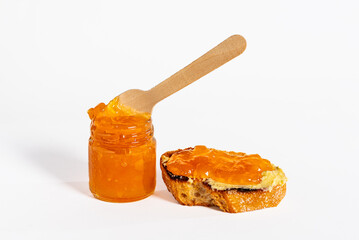 Glass jar with overflowing peach jam and a wooden spoon inside the preserve, next to a slice of bread with butter and jam.