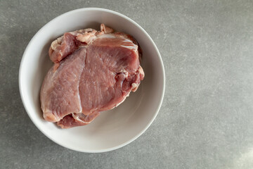 A piece of raw pork in a white bowl on a gray kitchen countertop.