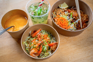 Vegetable salade with shrimp, pad thai, cucumber lemonade and coconut soup in paper bowls on bamboo pad on table.