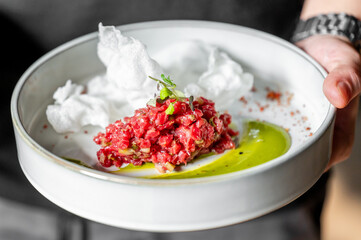 A gourmet dish featuring finely chopped raw meat topped with herbs, served on a white plate with a green puree and garnished with crispy elements, presented by a person's hand.