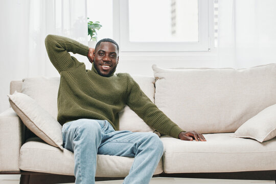 Relaxed young African man in a green sweater smiling cheerfully on a cozy sofa in a bright living room with natural light