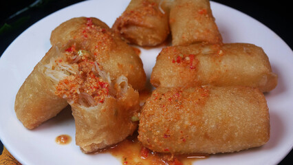 Close-up golden crispy risol or risoles kampung filled with vermicelli, coated in spicy chili sauce, served on a white plate. A savory and spicy Indonesian delicacy.