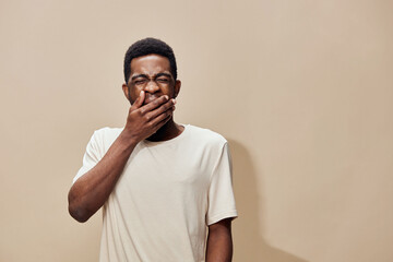 Young African man yawning with a funny expression, wearing a casual cream t shirt against a beige background, conveying tiredness and relaxation