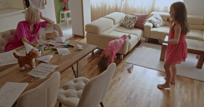 A mother watches her two daughters play and do cartwheels in their living room. One daughter does a cartwheel and the other dances around her.