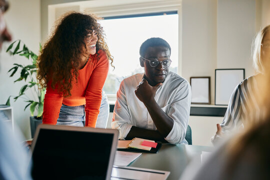 Diverse team engaged in a vibrant collaborative office meeting. Behind him stands a colleague, leaning over the table.