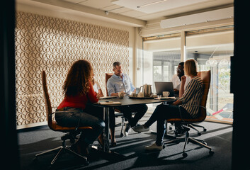 Diverse business team engaged in a productive office meeting discussion. Sunlight streams in through the large window behind them.
