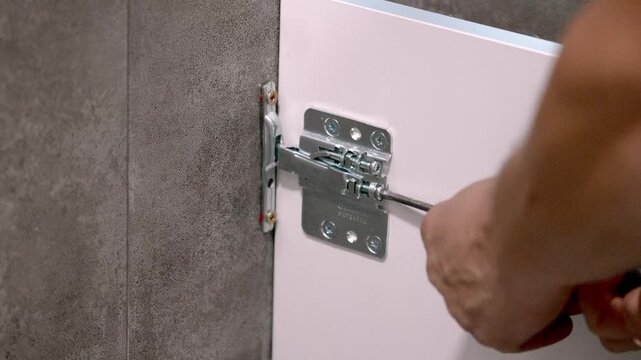 Worker tightening hinge screw on white cabinet door, focusing on precise installation against gray tiled wall background, showcasing professional home improvement craftsmanship