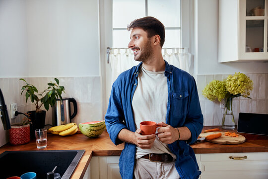 Smiling man in kitchen holding cup looking out the window - Powered by Adobe