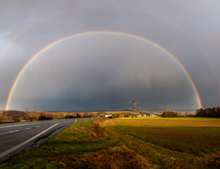 A brilliant full-arc rainbow stretches across a rural landscape, with a faint secondary rainbow...