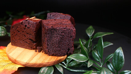 Tempting premium brownies, dark chocolate in color with crunchy almond topping slices, showing the details and texture of the soft cake, looks elegant with artificial leaves and a dark background.