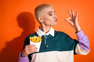 Stylish young woman enjoying french fries, displaying a cheerful expression, against a vibrant orange background