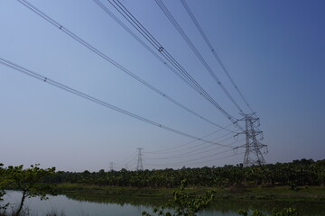 High-voltage power lines supported by transmission towers traversing a landscape with trees and water bodies, likely part of an electrical grid system