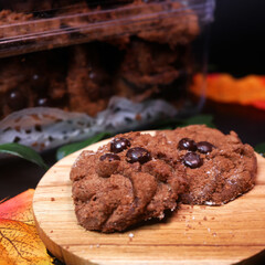 Crispy chocolate cookies with choco chips on a wooden plate, a cookie-filled jar in the background, greenery, and a warm atmosphere, perfect for Eid treats.