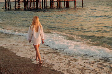 Young female in white shirt exploring coastline at sunset. Warm tones, calm sea waves, and carefree vacation spirit. Perfect for holiday promotions, coastal living, and relaxation themes.